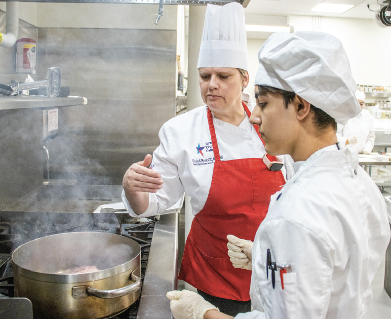 SE Campus culinary coordinator and instructor Katrina Warner helps SE student Gabriel Ortiz with placing a rack of lamb into a pot to cook for his assignment.